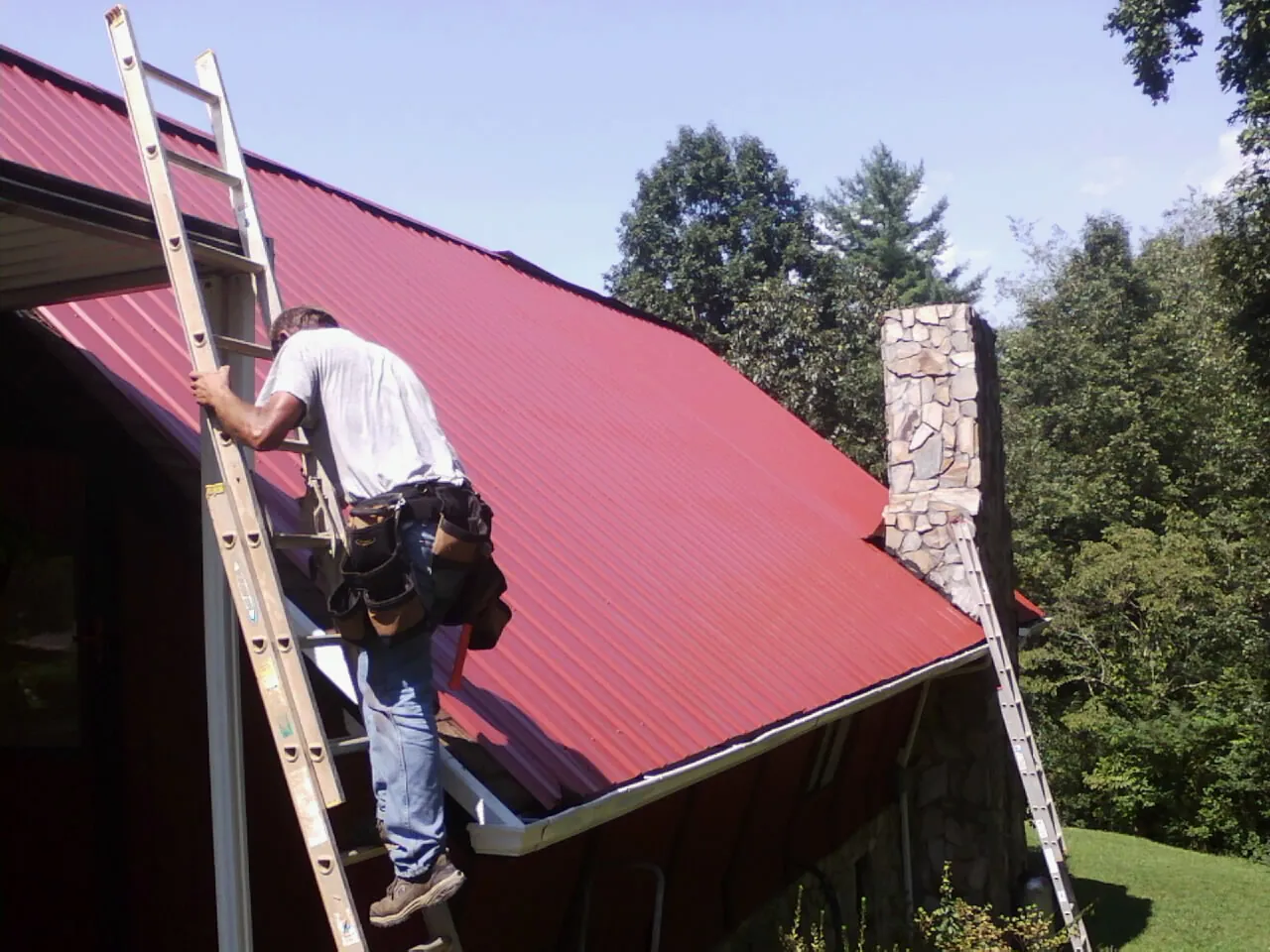 Roofer on a ladder beside a red metal roof and stone chimney — Soffit & Fascia Repair by Roofing Brothers in Hackettstown